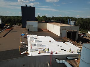 Workers in safety gear install white insulated rooftop panels on a flat industrial roof near a dark blue vent tower.