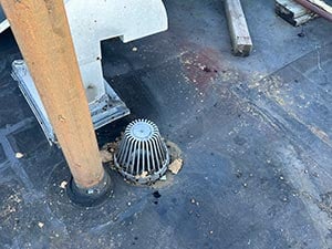 Flat-Roof-Repair-Post-Falls-ID-Idaho-2 | Flag Ship Foam & Coatings Rooftop surface with a rusted vertical support and a small circular metal roof drain/grate, surrounded by dark staining and debris in the area.